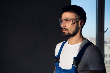 Mavi tulumlu bir erkek işçi gölgede duruyor.