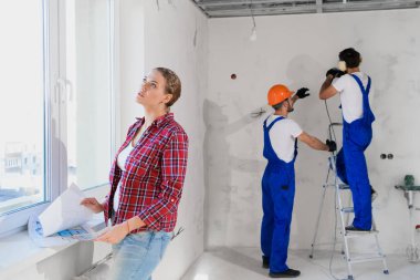 Kadın pencerede duruyor ve dairenin düzenini inceliyor. İnşaatçılar arka planda çalışıyor.