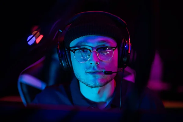 A young esports player sits at a computer during an online game tournament. Neon lighting of the esports arena. A hat and headphones with a headset. — Stock Photo, Image