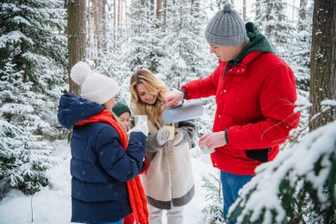 Anne ve baba çocuklarla birlikte karla kaplı bir ormanda çam ağaçlarıyla kışın tadını çıkarmak için dışarı çıktılar. Termosta lezzetli bir sıcak çay..