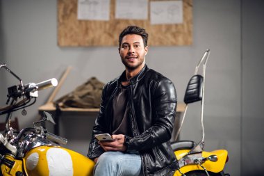 Garajda deri ceketli genç bir motorcu bir fotoğraf ve blogu için gülümsüyor..
