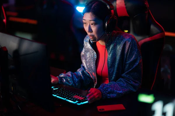 A young asian gamer wearing a headset and headphones on her head trains with a team of cyber athletes in a bootcamp. Neon light, blurry background. — Stock Photo, Image