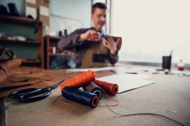 Bir terzinin masaüstündeki renkli ipliğin birkaç makarası, arka planda odaksız bir şekilde deri bir ürün dikiyor.
