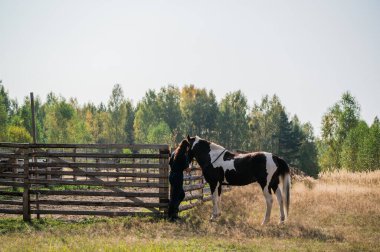 Genç seyis kız çitlere beyaz benekli kahverengi bir at bağlıyor.