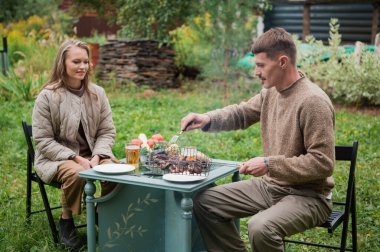 Bir adam, piknik sırasında karısına taze ızgara et tattırır. Köyün yaşam tarzı. Lezzetli yemekler, ızgara sebzeler ve birayla dolu bir masa..