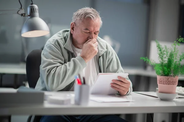 Under coronavirus pandemin sitter en vuxen man på ett kontor vid ett skrivbord, läser dokument och har en rinnande näsa — Stockfoto