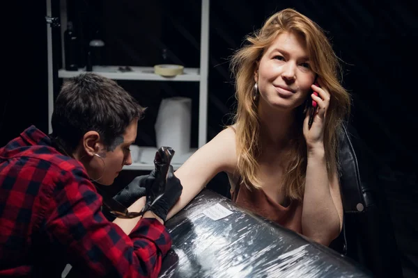 Un'esperta tatuatrice con un viso tatuato riempie un tatuaggio al suo cliente una ragazza con i capelli rossi mentre parla su uno smartphone. — Foto Stock