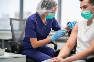 Klinikte maske ve eldiven takan deneyimli bir laboratuvar asistanı genç bir adamı aşılıyor.