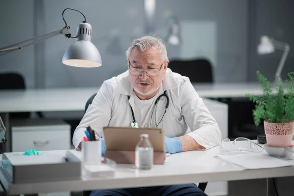 Een ervaren grijsharige arts voert een online consult met de patiënt uit via een videolink op een tablet. E-gezondheidsconcept — Stockfoto