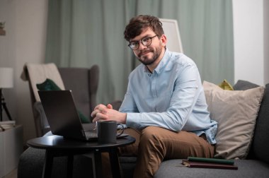 Gözlüklü sakallı bir adam evde çalışıyor, bir sehpanın önünde elinde dizüstü bilgisayar ve bir fincan kahve ile oturuyor.