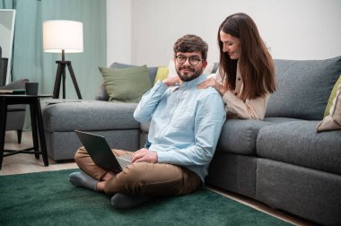 Bakıcı kadın kocasına omuz masajı yaparken o da evde dizüstü bilgisayarda çalışıyor ve yerde oturuyor.