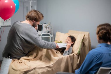 Bir ağabey küçük bir kıza hediye veriyor, kız yatakta yatıyor, bütün aile doğum gününde onu hastanede ziyarete gelmiş.