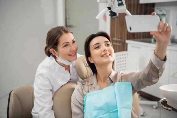 Joven médico hermoso, dentista, higienista u ortodoncista y su paciente tomando selfie con un teléfono inteligente después de un tratamiento exitoso — Foto de Stock