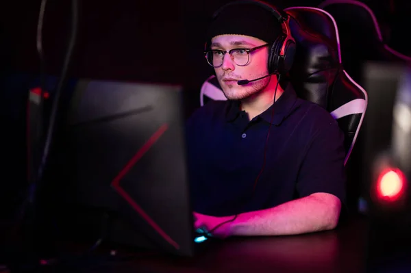 A young esports player sits at a computer during an online game tournament. Neon lighting of the esports arena. A hat and headphones with a headset. — Stock Photo, Image