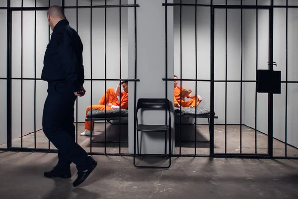 Un guardia de la prisión con un bastón hace un recorrido por las celdas de la prisión antes de apagar las luces. Dos prisioneros con túnicas rojas están tras las rejas. —  Fotos de Stock