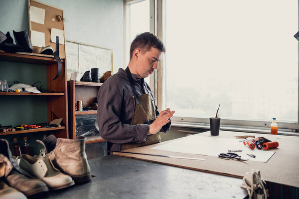 A young shoemaker makes a drawing for a pattern for leather shoes on a table in his workshop