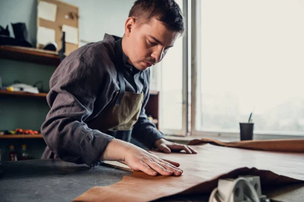 Before making shoes out of leather, a young guy lays them out on the table in the workshop