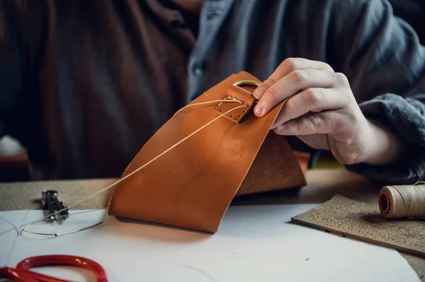 Sentado en una mesa en el taller un joven cose manualmente elementos de cuero el uno al otro — Foto de Stock