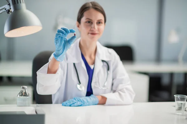 Uma jovem médica de luvas médicas azuis mostra a um paciente uma cápsula com um novo medicamento. Retrato no fundo do consultório médico — Fotografia de Stock