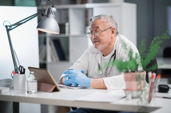 An elderly doctor sees patients online using a tablet and a video call. Remote assistance of a medical specialist. E-health concept