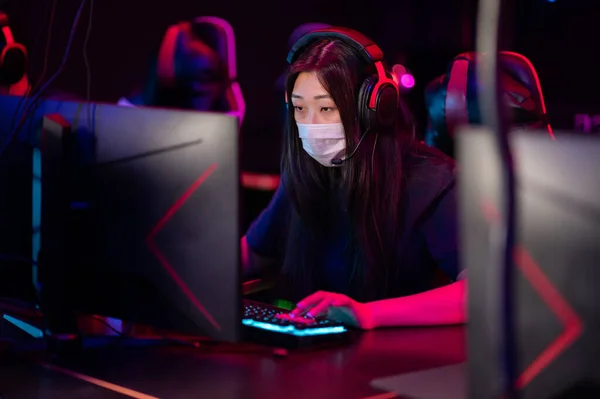 A close-up portrait of a gamer girl wearing headphones and mask in a computer club while playing a game — Stock Photo, Image