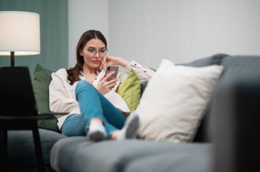 Gözlüklü, serbest çalışan güzel bir kız kanepe yastıklarına uzanıyor, elinde bir telefon tutuyor ve arkadaşlarıyla iletişim kuruyor.