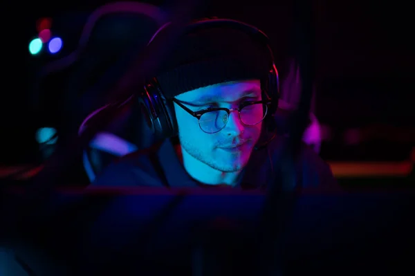 Un giovane giocatore esporta si siede a un computer durante un torneo di gioco online. Illuminazione al neon dell'arena degli sport. Un cappello e delle cuffie con un auricolare. — Foto Stock