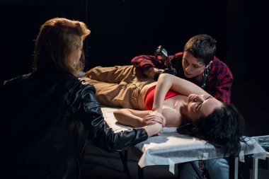 Genç ve güzel bir esmer, dövme salonunda yan tarafına dövme yaptırıyor. Kız arkadaşı yanına oturuyor ve elini tutuyor.