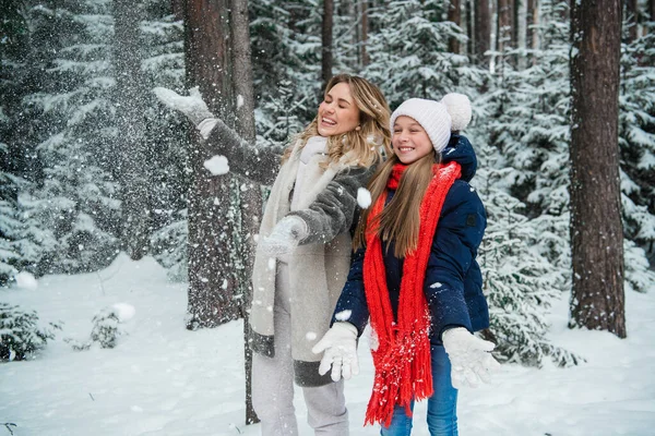 Uma jovem mulher bonita em um casaco de pele e uma menina bonita em uma jaqueta, chapéu e cachecol vomitar neve no inverno na floresta. — Fotografia de Stock