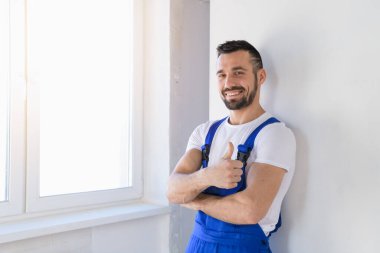 İşçi tulumu giymiş, baş parmağını gösteriyor ve gülümsüyor.