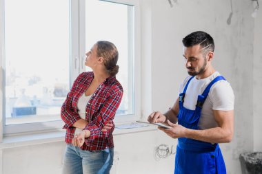 İnşaatçı, kadınla birlikte daireyi kontrol eder ve bir deftere yazar.