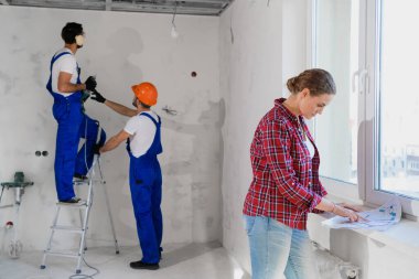 Bir müşteri, arka planda, bir apartman dairesi planlamasını inceliyor. Tulumlu adamlar, elinde matkapla merdivende duruyorlar.