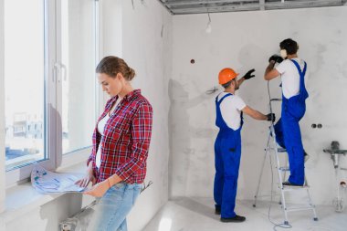 Kadın pencerede duruyor ve dairenin düzenini inceliyor. İşçiler arka planda çalışıyor.