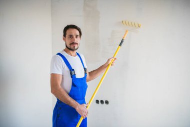 Tulumlu gülümseyen erkek ressam duvarın yanında duruyor ve elinde bir silindir tutuyor.