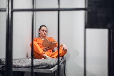 Dövmeli bir yüzü olan genç bir mahkum hücrede uzaktan eğitiliyor. Ranzada oturmuş ders kitabı okuyor..