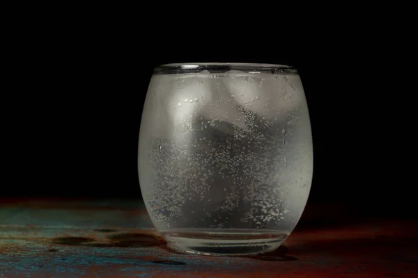 Glass of ice water filled with carbonated water on a dark background and rustic surface. Heat and thirst concept.