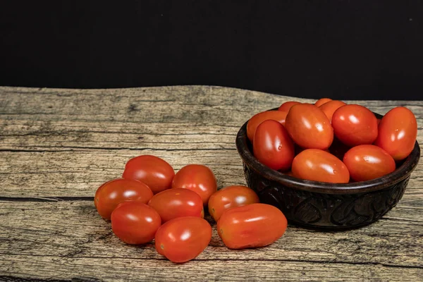Cherry tomatoes in an African ebony wood bowl on a wooden surface with a black background. Some cherry are fallen on the table. concept vegetable, cooking,