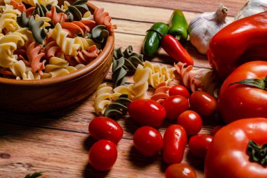 Üç renkli fusilli makarna ve kirazlı terracotta kasesi ve domates, kırmızı biber, sarımsak ve maydanoz. Yiyecek hammaddesi konsepti. 45 derece açı.