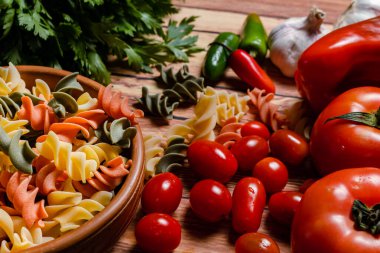 Üç renkli fusilli makarna ve kirazlı terracotta kasesi ve domates, kırmızı biber, sarımsak ve maydanoz. Yiyecek hammaddesi konsepti. 45 derece açı.