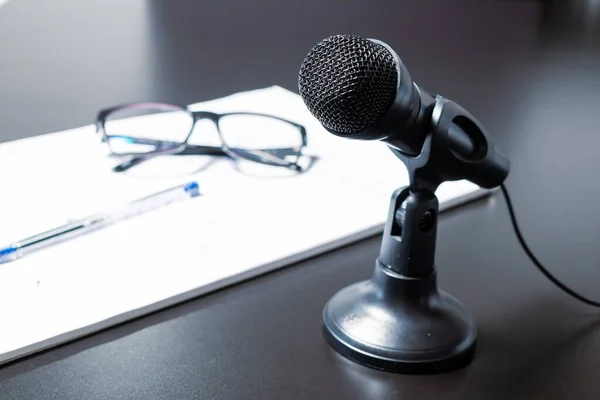 Small black desk microphone with cable and low stand on a black table next to notepad, glasses and glasses. Modern style, Communication and speech concept.