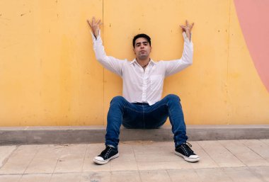 Kolları açık, sarı bir duvara yaslanmış genç Latin bir adam. Modern giysiler.