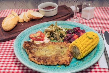 Etli şnitzel, domates salatası, yeşil fasulye, pancar kökü ve haşlanmış yumurta ve geleneksel bir otel veya restoran masasında mısır koçanı. Yatay görünüm