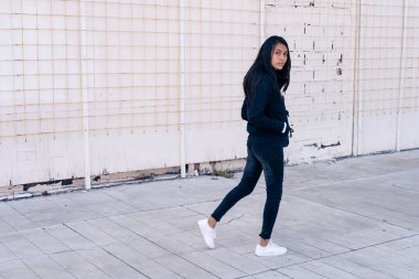 Siyah elbiseli genç bir Latin kadın yürüyor ve arkasına bakıyor. Ellerini cebine koy..