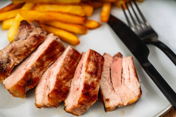 Grilled pork breast cut into strips on a plate with french fries on a table.