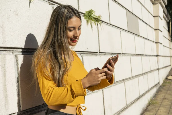 Portrait of a young Latina woman checking her smartphone or cell phone leaning against a wall. Technology and communications concept.