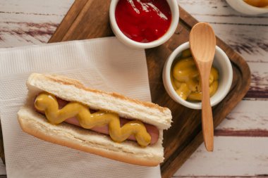 Top view of a hotdog with mustard on a wooden board with pots with ketchup and mustard. Fast food concept. clipart