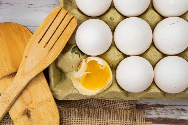 Normal view of an egg carton container with organic white chicken eggs with one broken egg on a rustic wooden countertop.