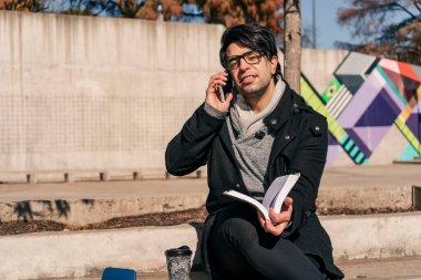 Adam halka açık bir parkta beton bir bankta oturmuş kitap okuyor ve cep telefonuyla konuşuyor. İletişim ve bilgi kavramı.