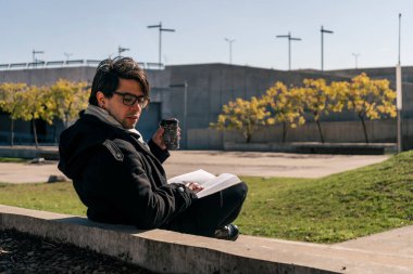 Adam halka açık bir parkta beton bir bankta oturmuş kitap okuyor ve güneşin altında sıcak bir şeyler içiyor..