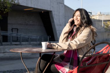 Güneşli bir günde bir kafede kahve içerken cep telefonuyla konuşan güzel Latin bir kadın. Rahatlama ve boş zaman, iletişim, teknoloji konsepti.